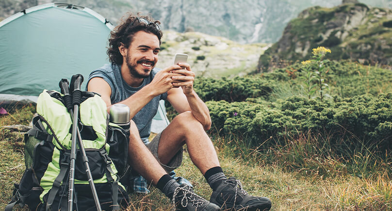 Junger Mann sitzt mit seinem Wanderrucksack vor einem Zelt und nutzt sein Smartphone